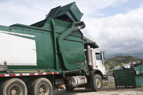 A large green commercial waste collection truck with a white cab is parked outdoors on a gravel surface, with its loading compartment raised and tilted to empty its contents. The truck's textured surface shows signs of wear typical of heavy-duty refuse vehicles, with visible hydraulic mechanisms and metal panels. In the background, a green waste bin filled with mixed rubbish, including plastic bags and other refuse materials, is positioned on the right side of the image. The setting appears to be an open area, possibly a waste collection site near Penge, with distant hilly landscape and partly cloudy sky overhead. The scene reflects a routine rubbish collection operation carried out by Business Waste Removal Penge, illustrating the practical aspect of commercial waste disposal services in the local area. The overall lighting is natural, highlighting the metallic and plastic textures of the equipment and waste containers, emphasizing cleanliness and operational detail relevant for waste management and rubbish removal services in the Penge region.
