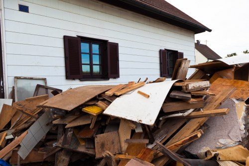 A large pile of assorted construction and demolition waste located on the paved driveway of a residential property, with a white exterior wall and dark brown window shutters in the background. The waste includes various wooden planks, particleboard panels, and plywood sheets, some with visible nails and rough edges. There are also a few pieces of drywall or plasterboard, with textured surfaces in white or off-white tones. The pile is situated adjacent to the house, occupying most of the visible driveway area, and the scene is illuminated with natural daylight, suggesting a clear or partly cloudy sky. The surrounding environment appears suburban, with the presence of neighboring houses partially visible in the distance. The image is relevant to rubbish removal services focusing on clearing construction or renovation debris, such as those provided by Business Waste Removal Penge, and emphasizes the scope of waste types managed in the local area.