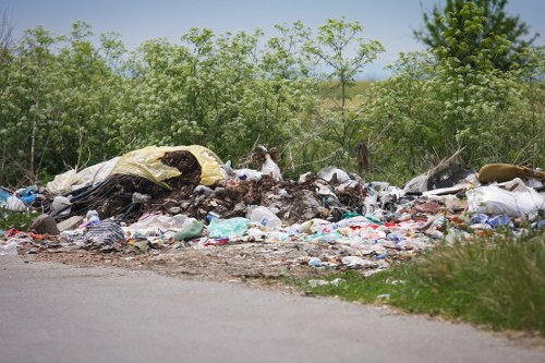A large pile of mixed waste materials is situated along the edge of a paved road, with trash including plastic bags, cardboard, and discarded fabric visible in the foreground. The waste is spread unevenly, with some items partially covered by dirt or pressed against each other, creating a cluttered scene. Behind the pile, there is a row of green bushes and small trees, with some taller trees further back, suggesting a rural or semi-rural environment. The sky above is clear with bright natural light illuminating the scene, highlighting the variety of textures and colors in the rubbish. This image exemplifies the type of roadside waste accumulation that professional rubbish removal services, such as those offered by Business Waste Removal Penge, often assist in managing, especially in areas near local towns or suburbs within the postcode region.