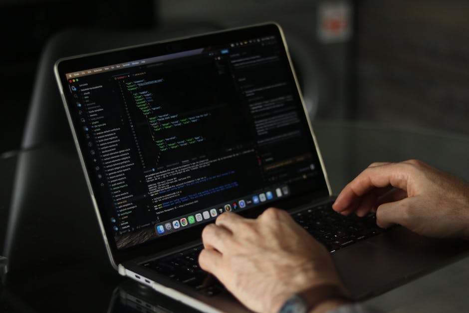 Close-up image of a person's right hand operating a laptop computer placed on a wooden desk surface. The hand is positioned over the keyboard, which features black keys with white lettering and backli
