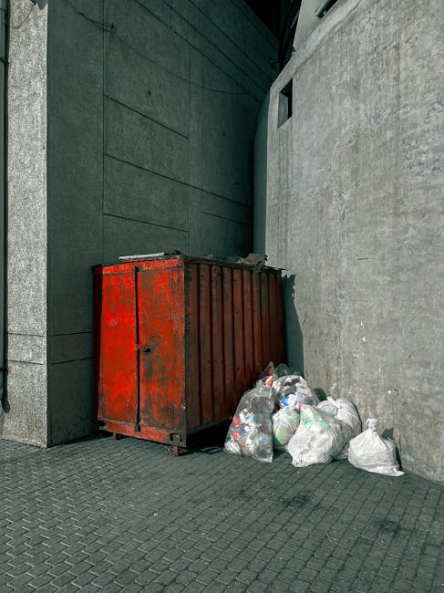 In the foreground, a large collection of mixed waste and rubbish is piled on a paved sidewalk near a metal railing, including cardboard boxes, paper bags, plastic packaging, and scattered loose items,